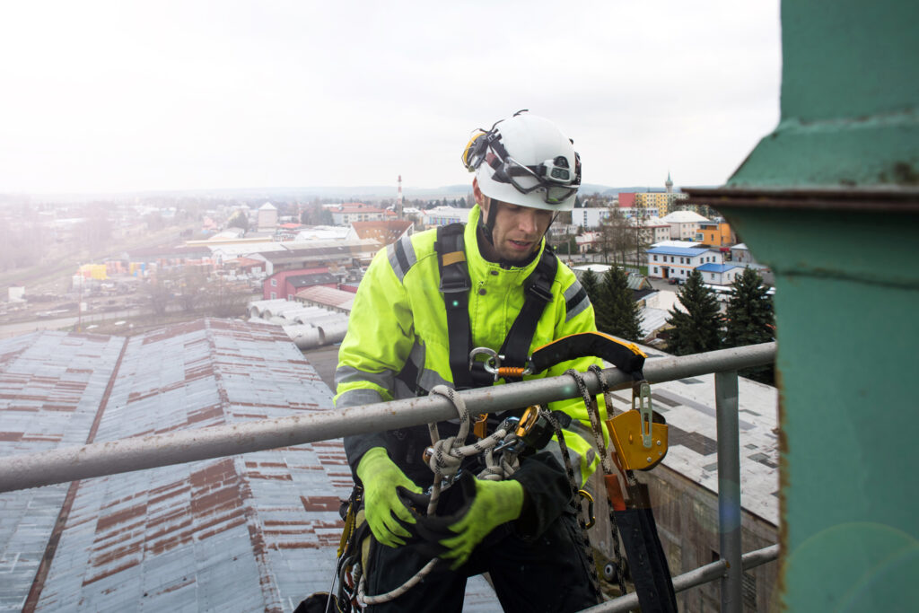 An industrial worker in high-visibility clothing and safety helmet meticulously secures himself with safety harnesses and ropes while working on a scaffold at a high elevation, against a backdrop of a cloudy sky and urban landscape, highlighting the paramount importance of adhering to safety protocols when working at heights.